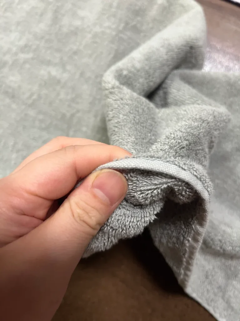 購入から数か月のグレーのスリムバスタオルを指でつまみ、厚みと柔らかさを見せる様子